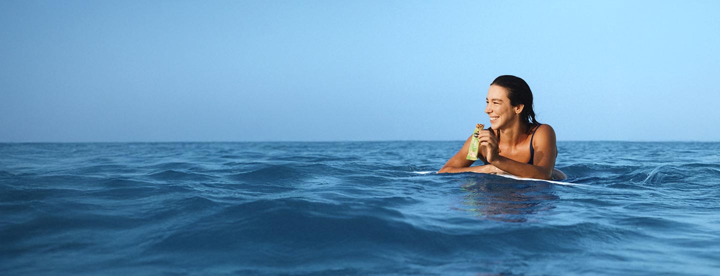 Marcela Witt no mar em cima de uma prancha de surf comendo barra biO2 Nuts Maça