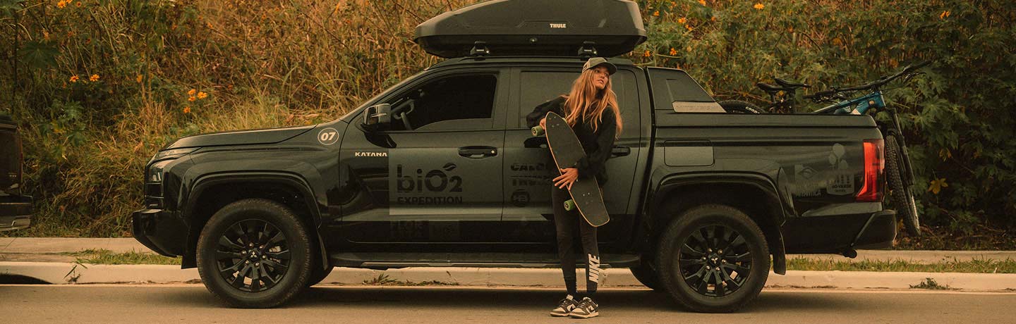 Menina loira com skate na mão na frente de uma Mitsubish L200 Katana com logo da biO2 na porta